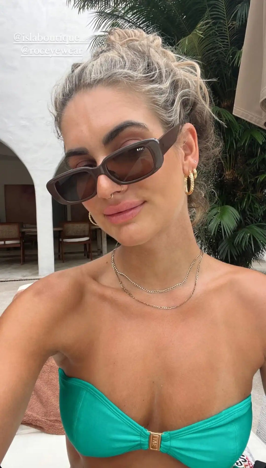 A woman with blonde, curly hair in a messy bun, wearing dark brown rectangular sunglasses, a gold nose ring, gold hoop earrings, two delicate gold chain necklaces, and a strapless teal bikini top with a gold clasp.