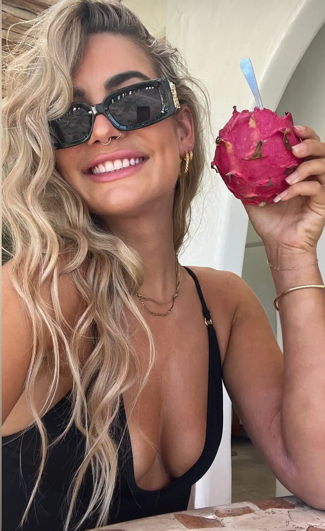 A smiling woman with long, wavy blonde hair and sunglasses holding a vibrant pink dragon fruit with a spoon.